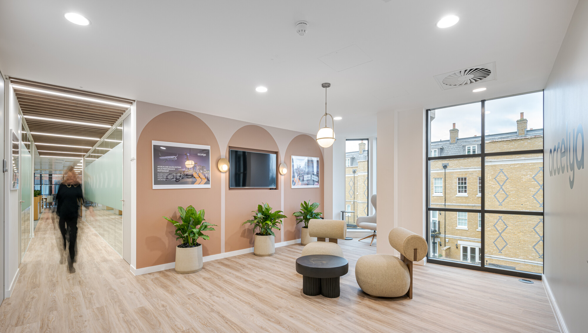 Accelya office designed by Two, featuring a lounge area with beige seating, potted plants, a black round table, and arched wall niches with mounted screens and lighting. A hallway with wooden flooring and frosted glass glazing is visible on the left.