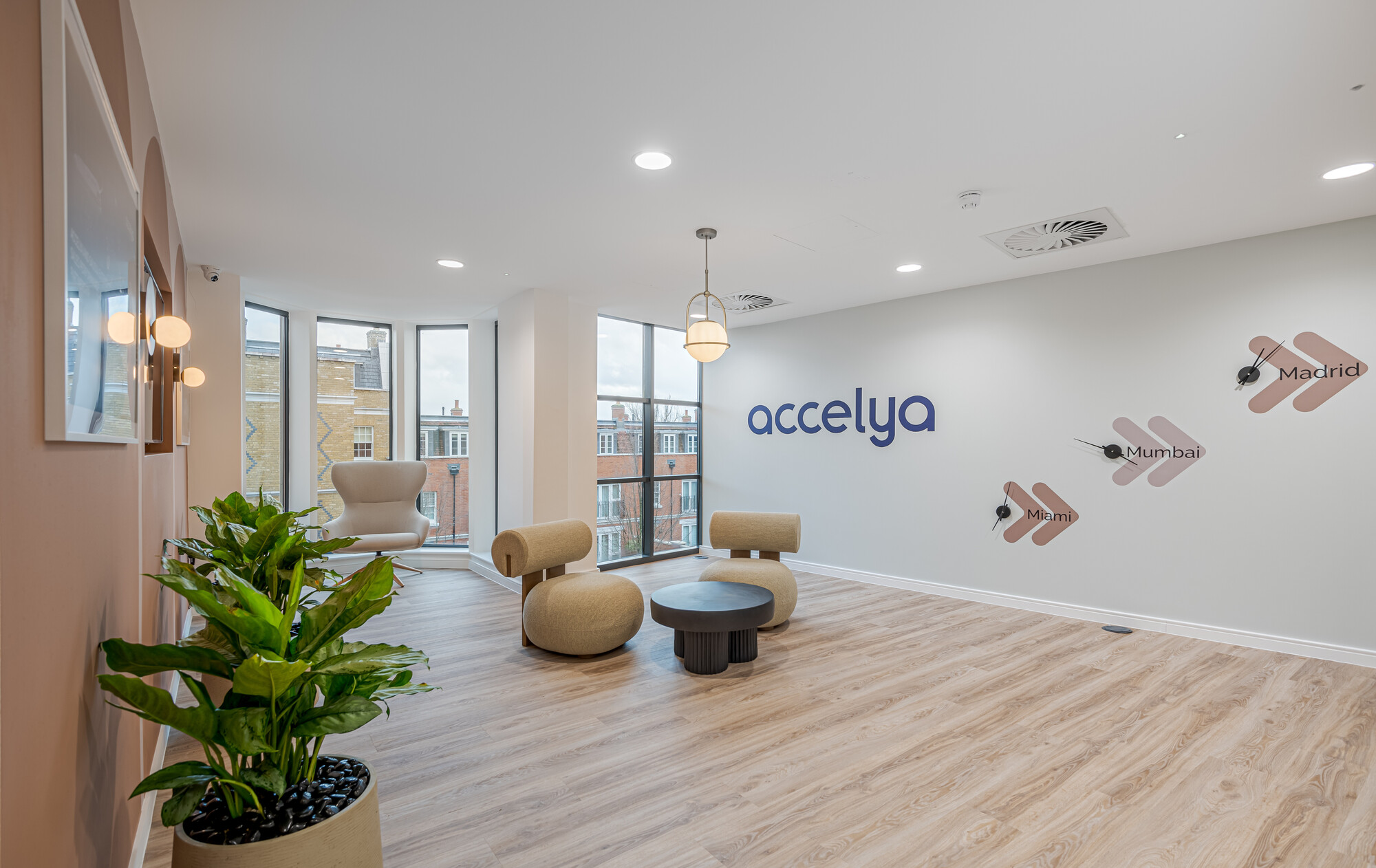 Accelya office designed by Two, featuring a lounge area with beige chairs, a black round table, and a light fixture. A wall displays the Accelya logo and clocks with city names, Miami, Mumbai, and Madrid. Potted plants and large windows with a view of brick buildings complete the space.