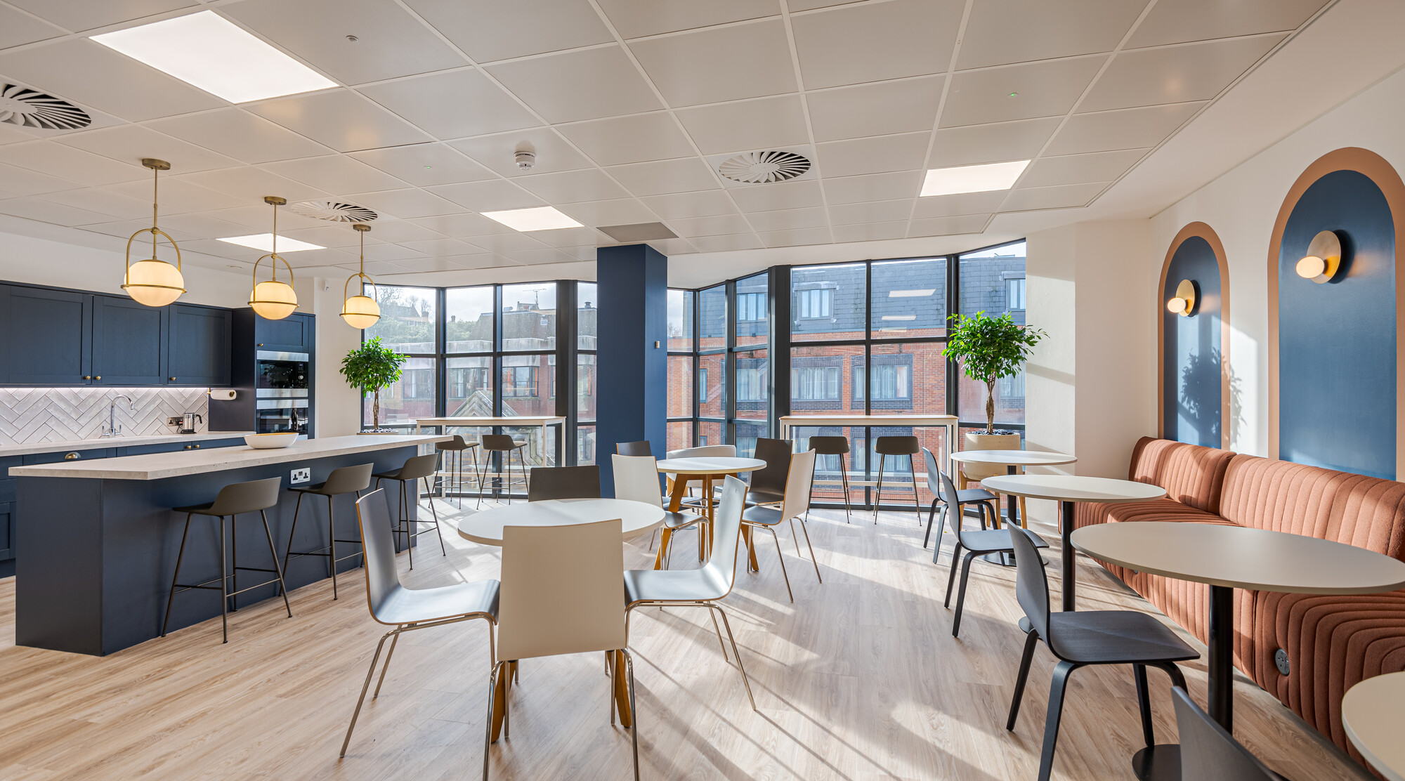 Accelya office designed by Two, featuring a kitchen and dining area with a navy blue island, herringbone-tiled backsplash, and brass pendant lights. The space includes round tables with chairs, a cushioned banquette with arched wall niches, potted plants, and large windows providing natural light and views of brick buildings.