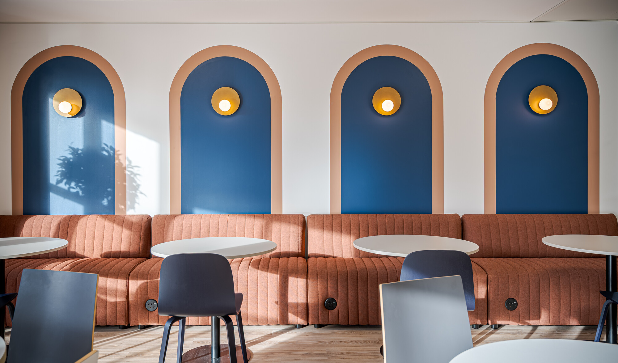 Accelya office designed by Two, showing a seating area with a rust-coloured ribbed banquette, round white tables, and navy blue chairs. The wall features arched panels in navy blue with soft beige borders, each adorned with a brass wall light. Sunlight casts a plant shadow on one of the panels.