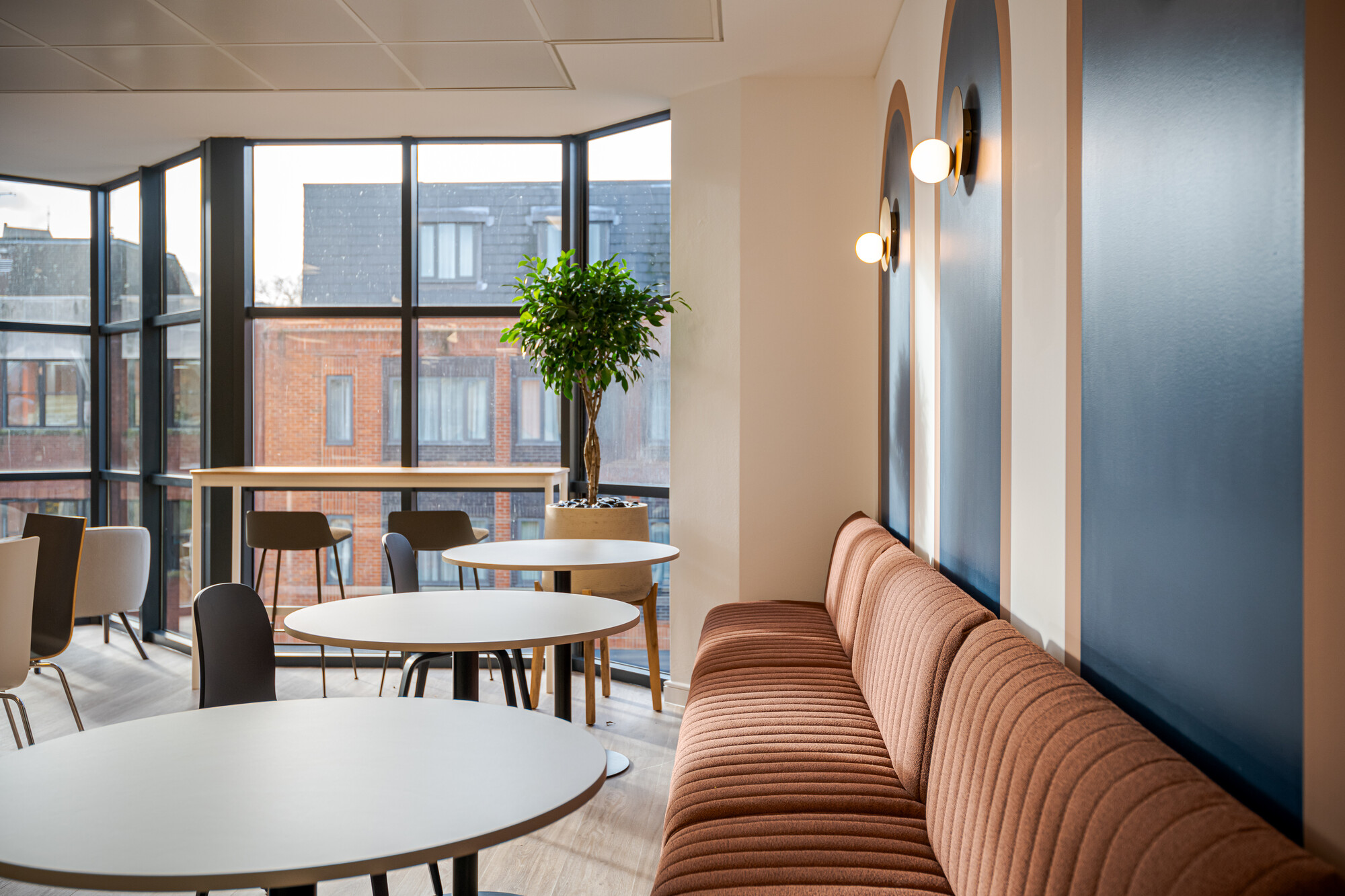 Accelya teapoint featuring a rust-coloured ribbed banquette, round white tables, and navy blue wall panels with brass sconces. Large windows provide views of red brick buildings, and the space includes a tall potted plant and a high table with bar stools.