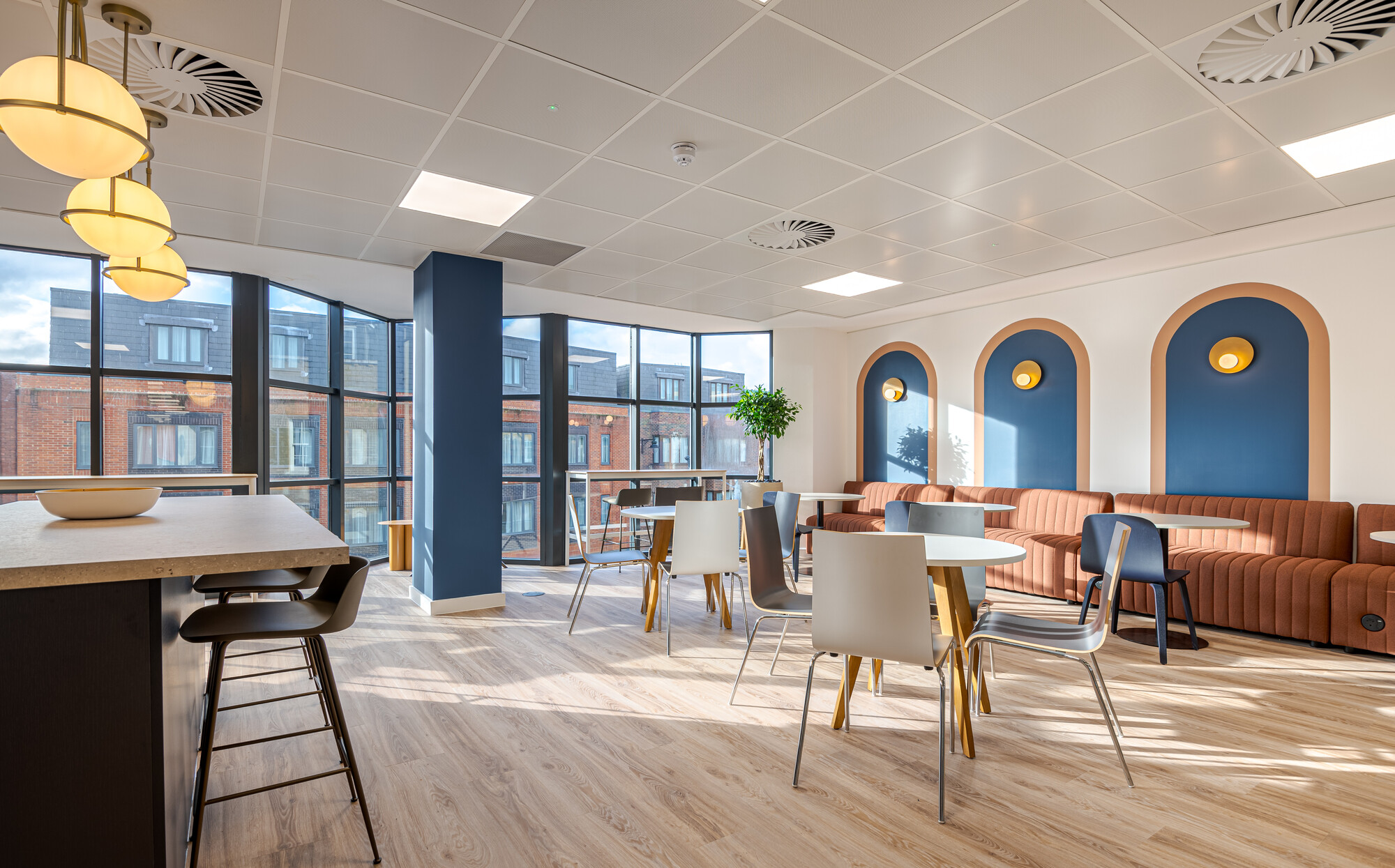 Accelya showcasing the breakout area with a central navy blue island, brass pendant lights, round tables, and chairs. The space includes rust-coloured ribbed banquette seating, arched navy blue wall panels with brass sconces, and large windows with views of red brick buildings. A tall potted plant adds greenery to the room