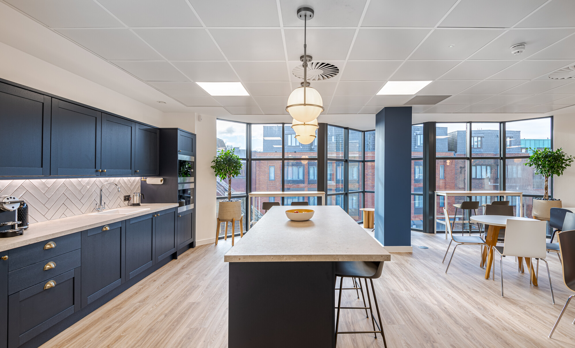 Teapoint in Accelya's office, a project delivered by Two. The space features navy cabinetry with brass handles, a herringbone-tiled backsplash, a large island with bar stools, and pendant lighting. The dining area includes round tables, chairs, potted plants, and floor-to-ceiling windows with views of brick buildings.