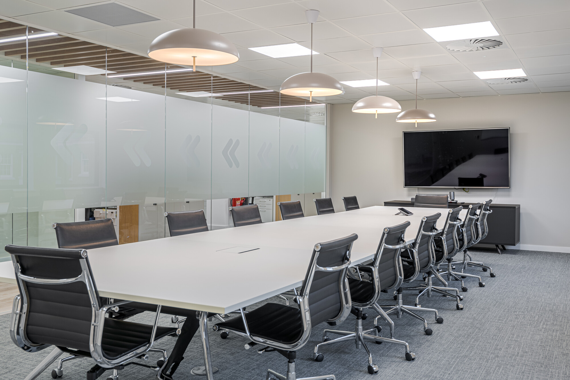 Large meeting room in Accelya's office by Two, with a long white conference table surrounded by black leather chairs on casters. Frosted glass walls with arrow motifs separate the space, and pendant lighting hangs overhead. A wall-mounted screen and a console unit complete the room.