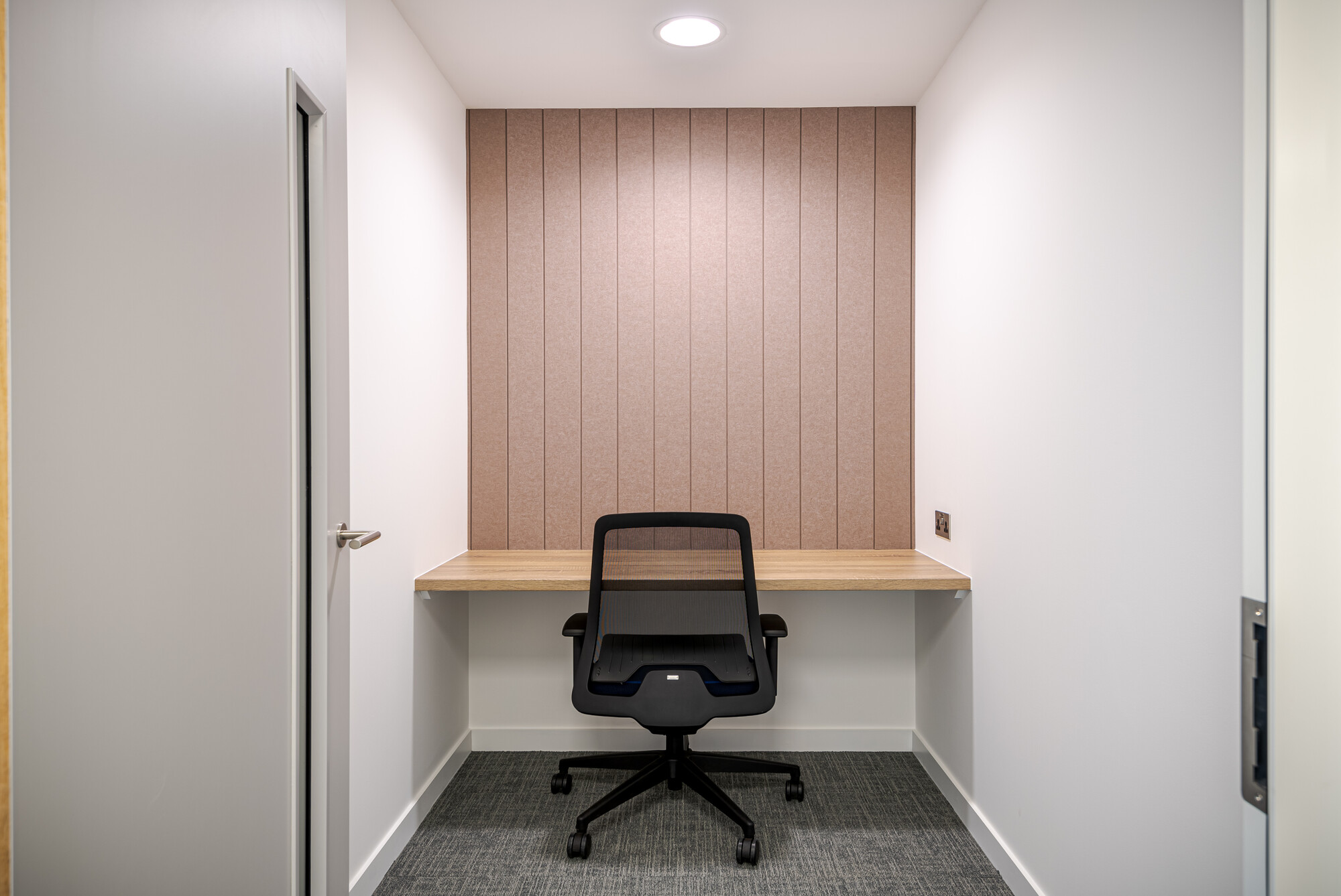 Privacy room in Accelya's office by Two, designed for individual use. The space includes a built-in wooden desk, a black mesh office chair, and a textured wall panel in a neutral tone. Simple and functional with a quiet atmosphere for focused work or virtual meetings