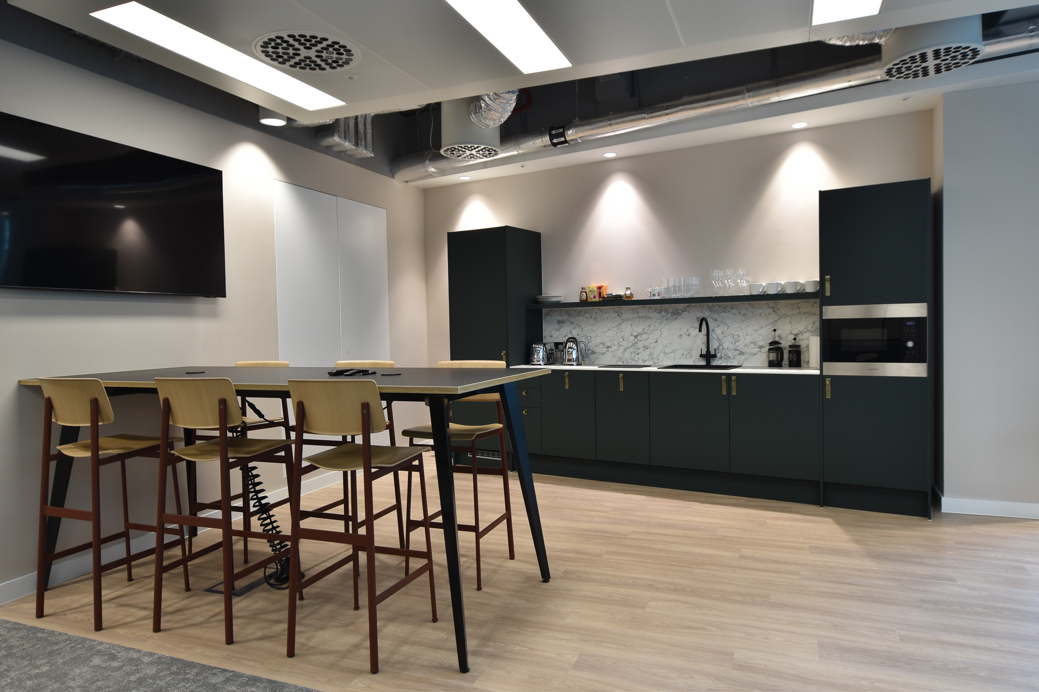 Casual and inviting kitchen area in Landsec Nova North office designed by Two, featuring bar stools and a green cabinet kitchen with marble countertops.