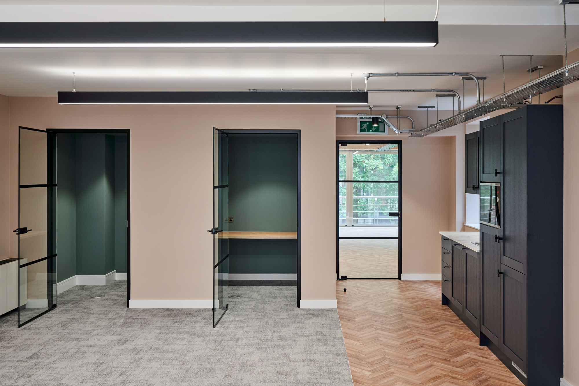 Nolan Partners office designed by Two, showing open glass doors leading to small rooms with dark green walls. The flooring transitions from grey carpet to herringbone wood to separate the different areas in the office with a dark kitchen area on the right.