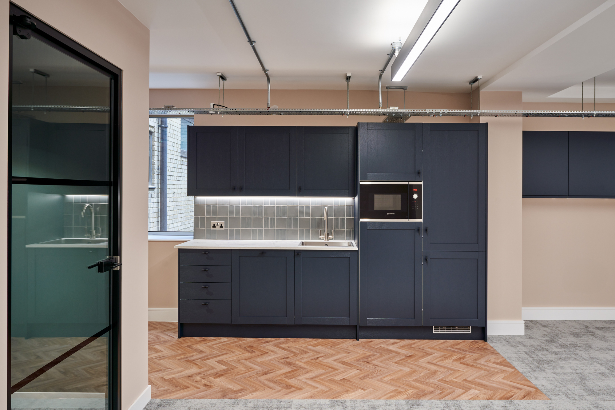 Kitchen area in Nolan Partners office by Two, with navy cabinetry, a tiled backsplash, and herringbone wood flooring adjacent to grey carpet.