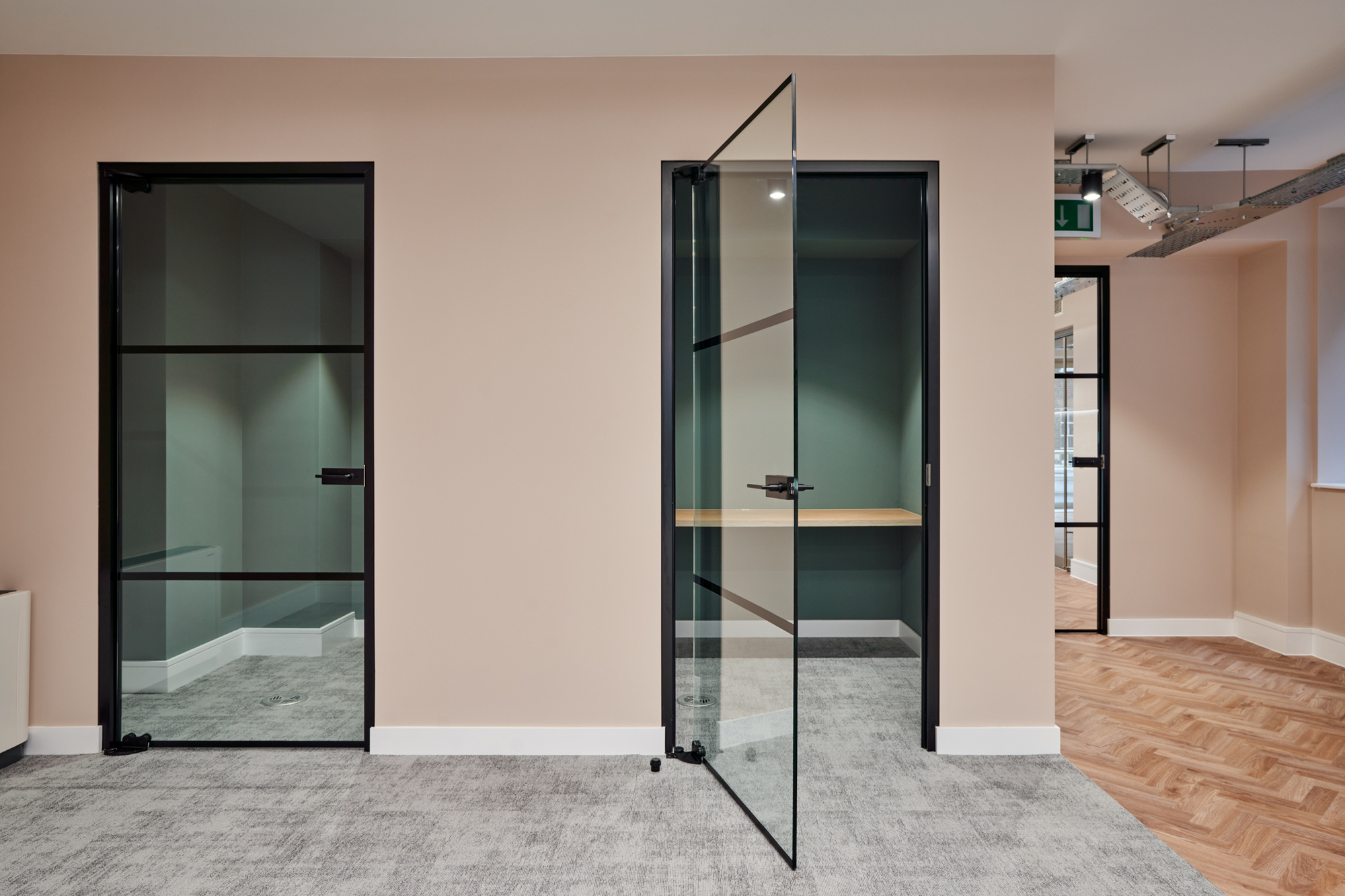 Glass doors with black frames in Nolan Partners office designed by Two, revealing two bespoke focus booths with a green feature wall and wood flooring.