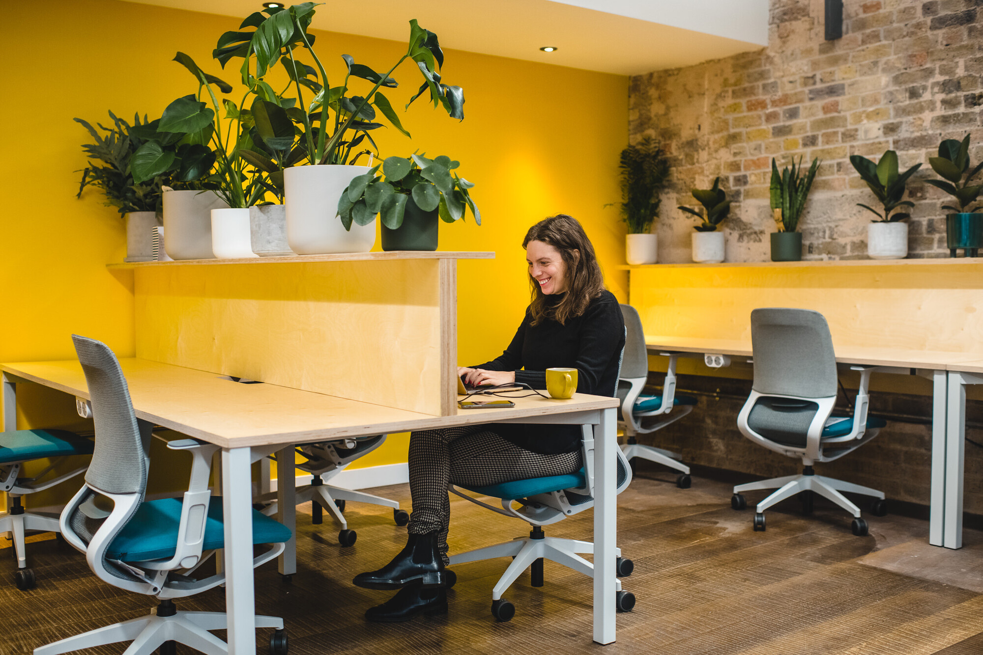 Bright workspace at Patch designed by Two, featuring a yellow wall, natural wood desks, and greenery, creating a welcoming atmosphere for co-working.