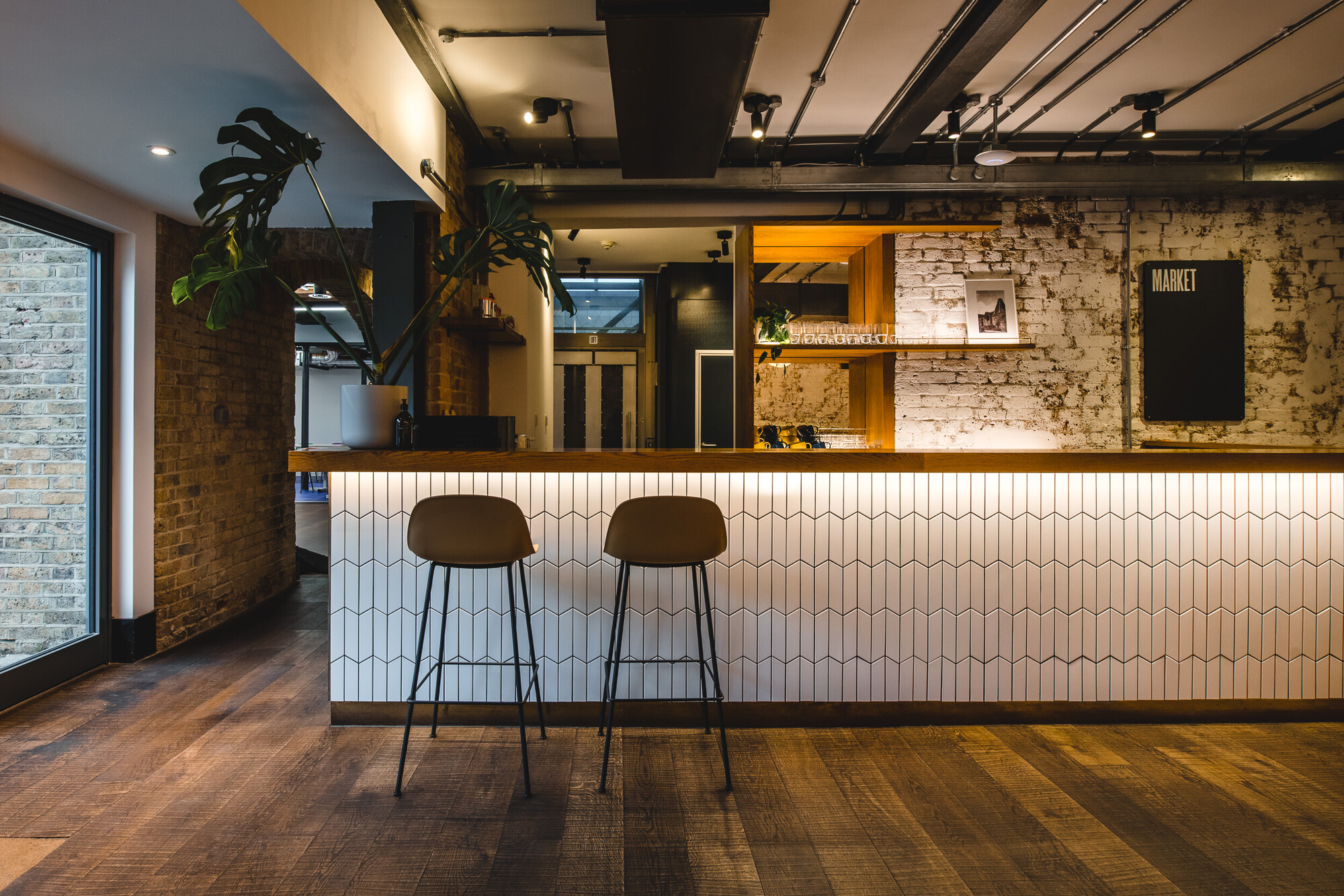 Patch office designed by Two showcasing a warm, industrial-style café area with a tiled counter, exposed brick walls, wooden flooring, and modern bar stools.