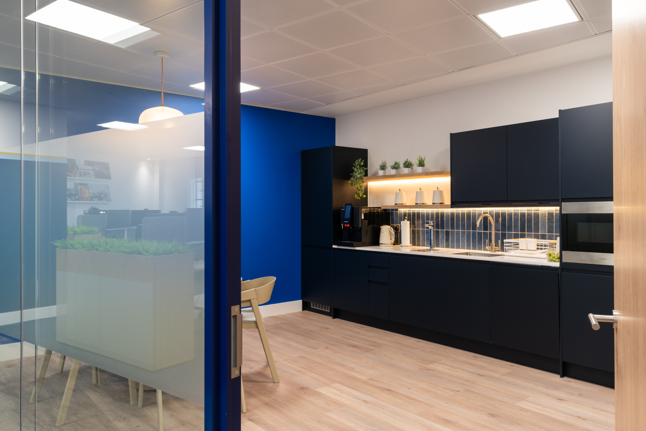 View of the teapoint area at the Port of London Authority office by Two, with dark cabinetry, a blue accent wall, and natural light filtering through glazing.