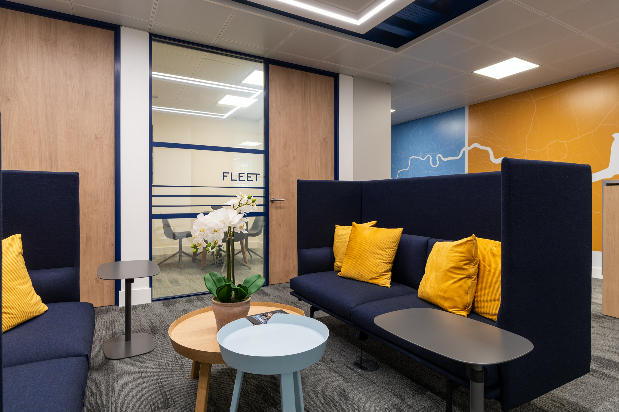 Informal seating area at the Port of London Authority office by Two, featuring navy high-back sofas, yellow cushions, and a river-themed mural.