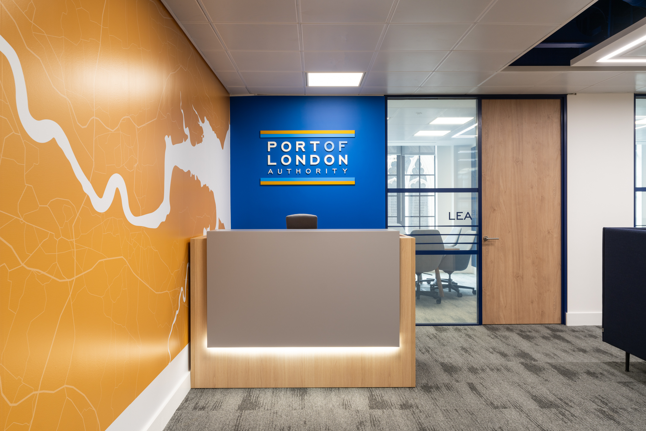 Reception area at the Port of London Authority office by Two, featuring a branded wall with river graphics, a sleek desk, and glass meeting room doors.