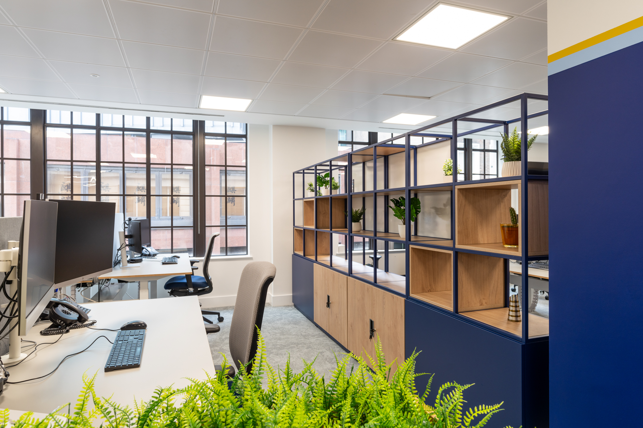 Port of London Authority office by Two, showcasing partitioned desks, navy shelving with natural wood accents, and greenery under large windows.