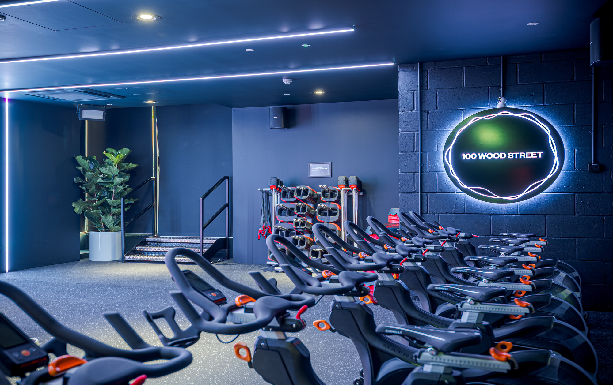 Indoor cycling area at ReFIT Gym, 100 Wood Street, designed by Two, with neon signage and rows of spin bikes under dynamic lighting.