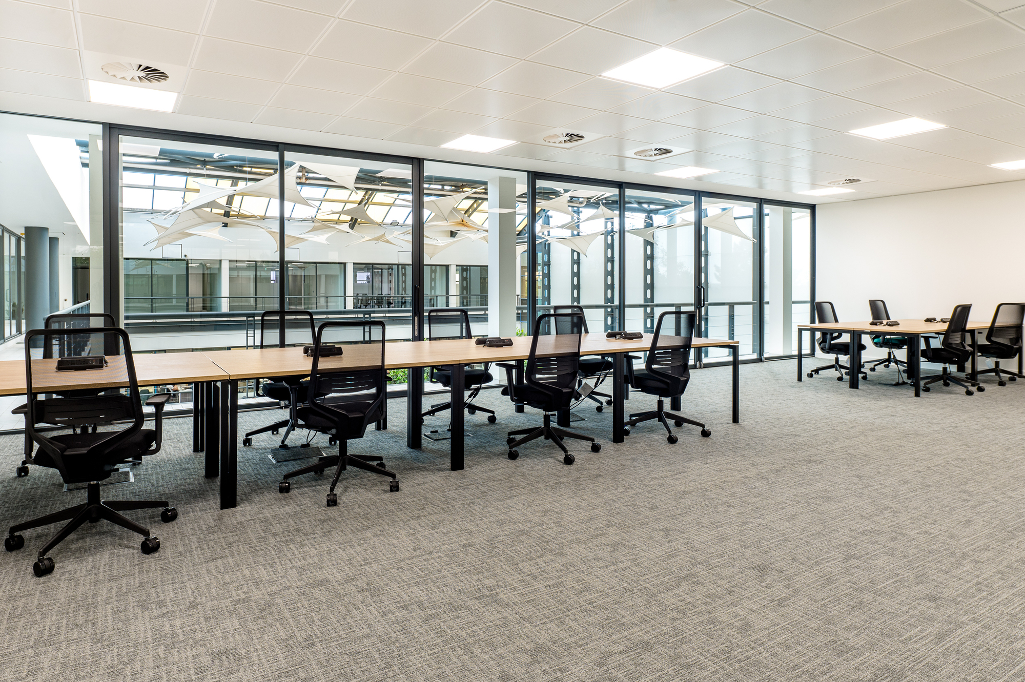 Bright workspace featuring large shared desks and ergonomic chairs, with floor-to-ceiling glass walls overlooking an atrium with architectural sails.