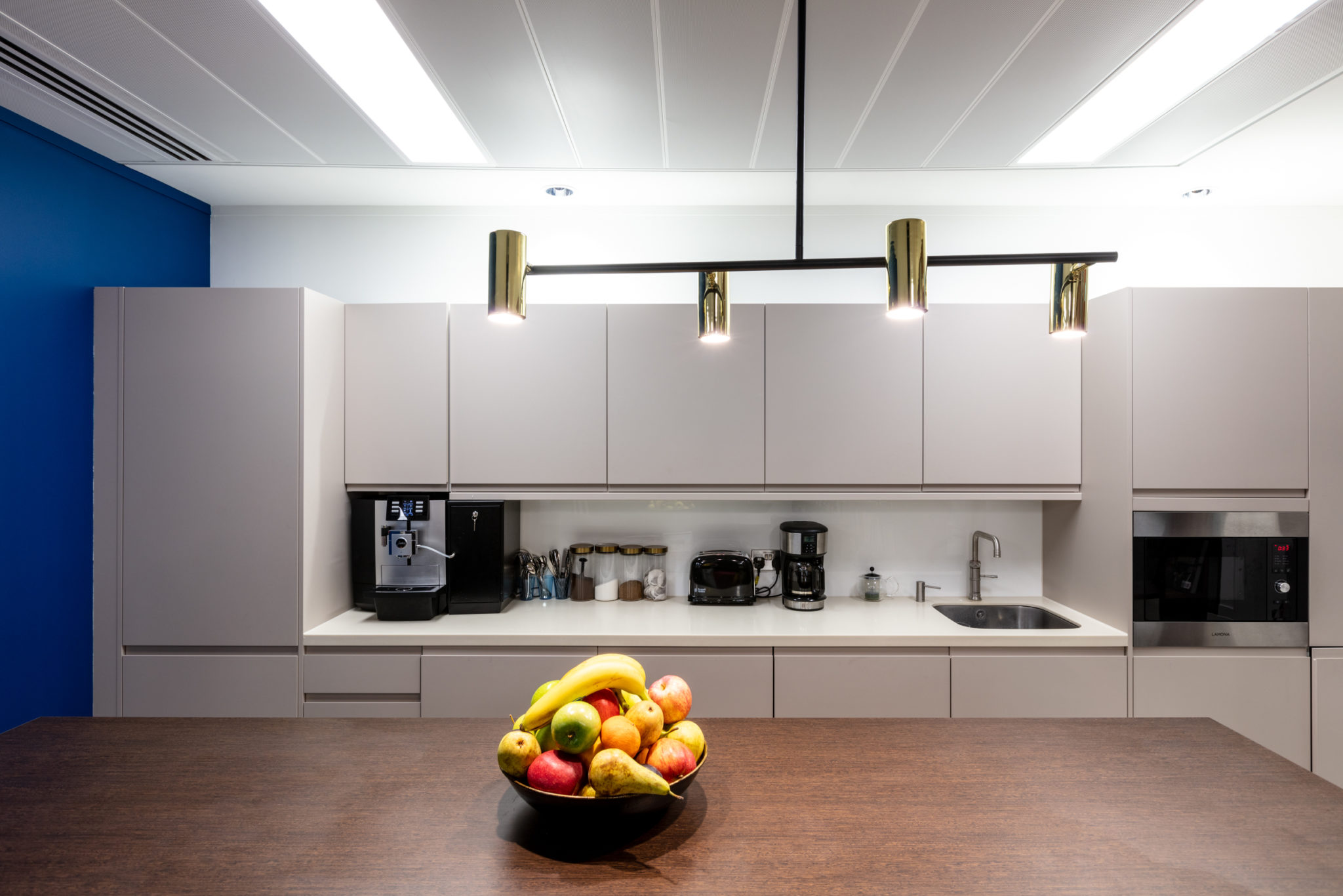 Contemporary tea point in the Urban Exposure workspace by Two, showcasing sleek cabinetry, modern appliances, and a fruit bowl centerpiece for a welcoming environment