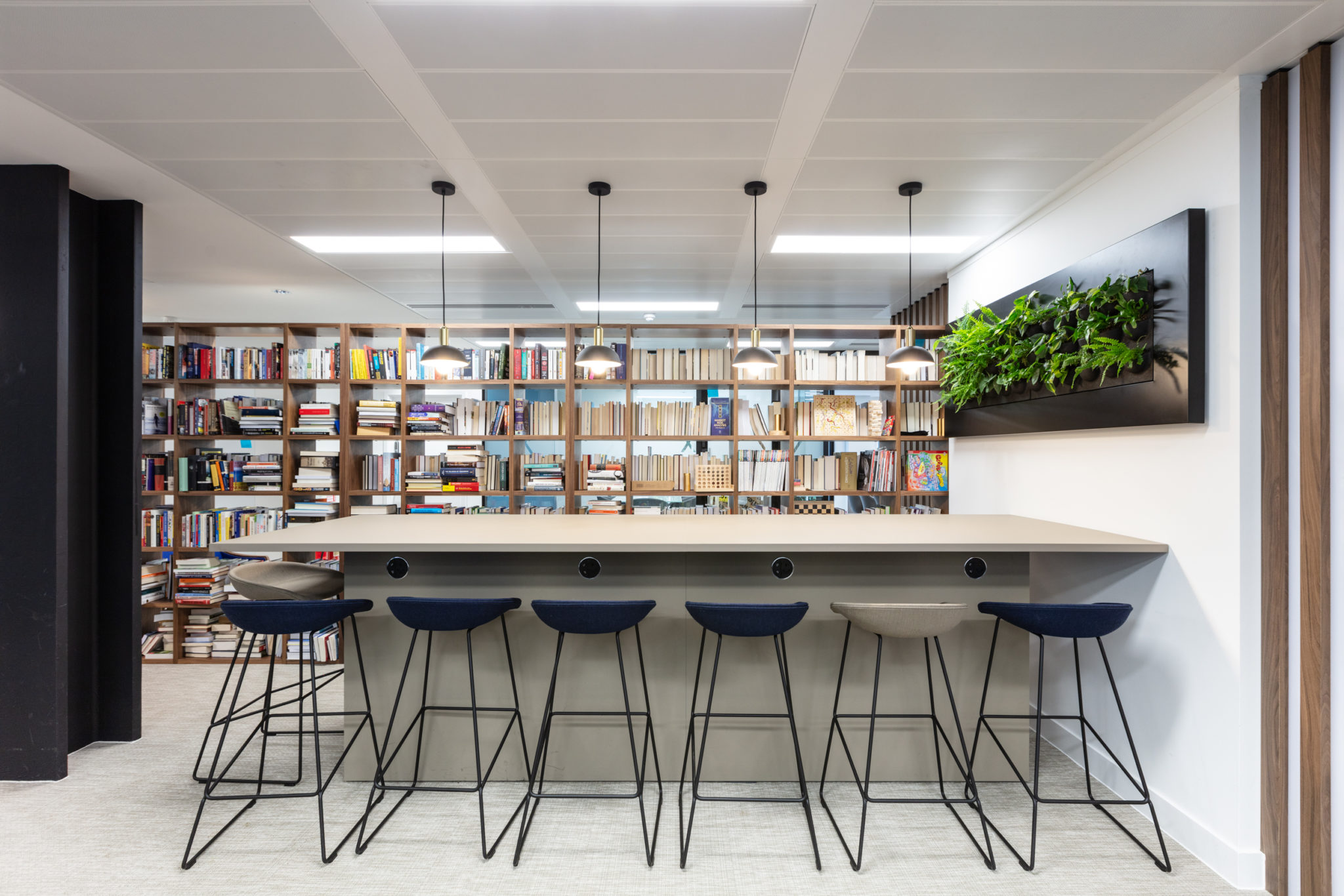 Urban Exposure workspace library by Two, showcasing a modern bar-height desk with integrated charging points, sleek stools, and lush greenery for a functional yet inviting setting.