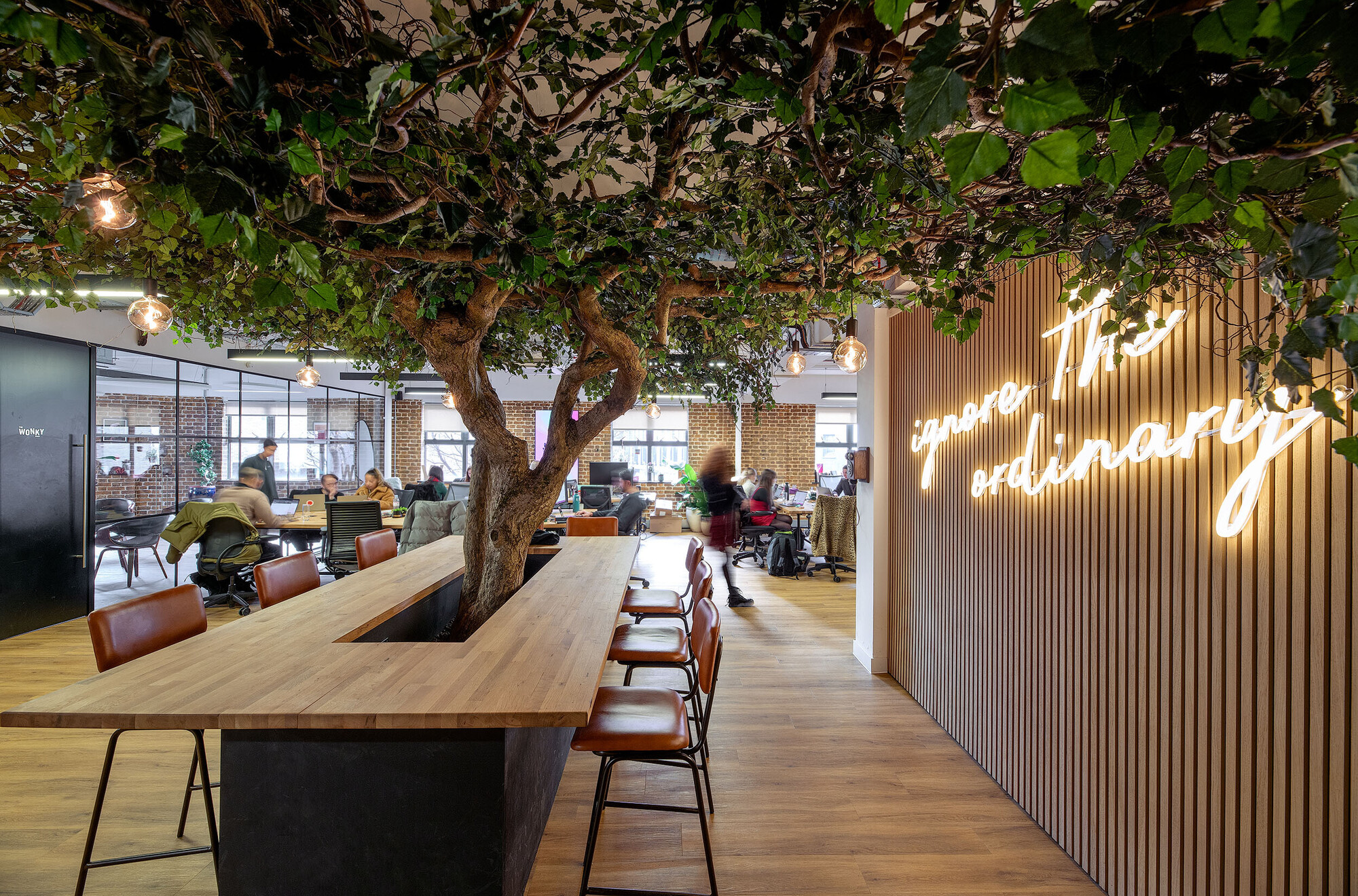 Wiser workspace designed by Two, featuring a central collaboration table with a striking tree centrepiece and a neon-lit wall reading 'ignore the ordinary.