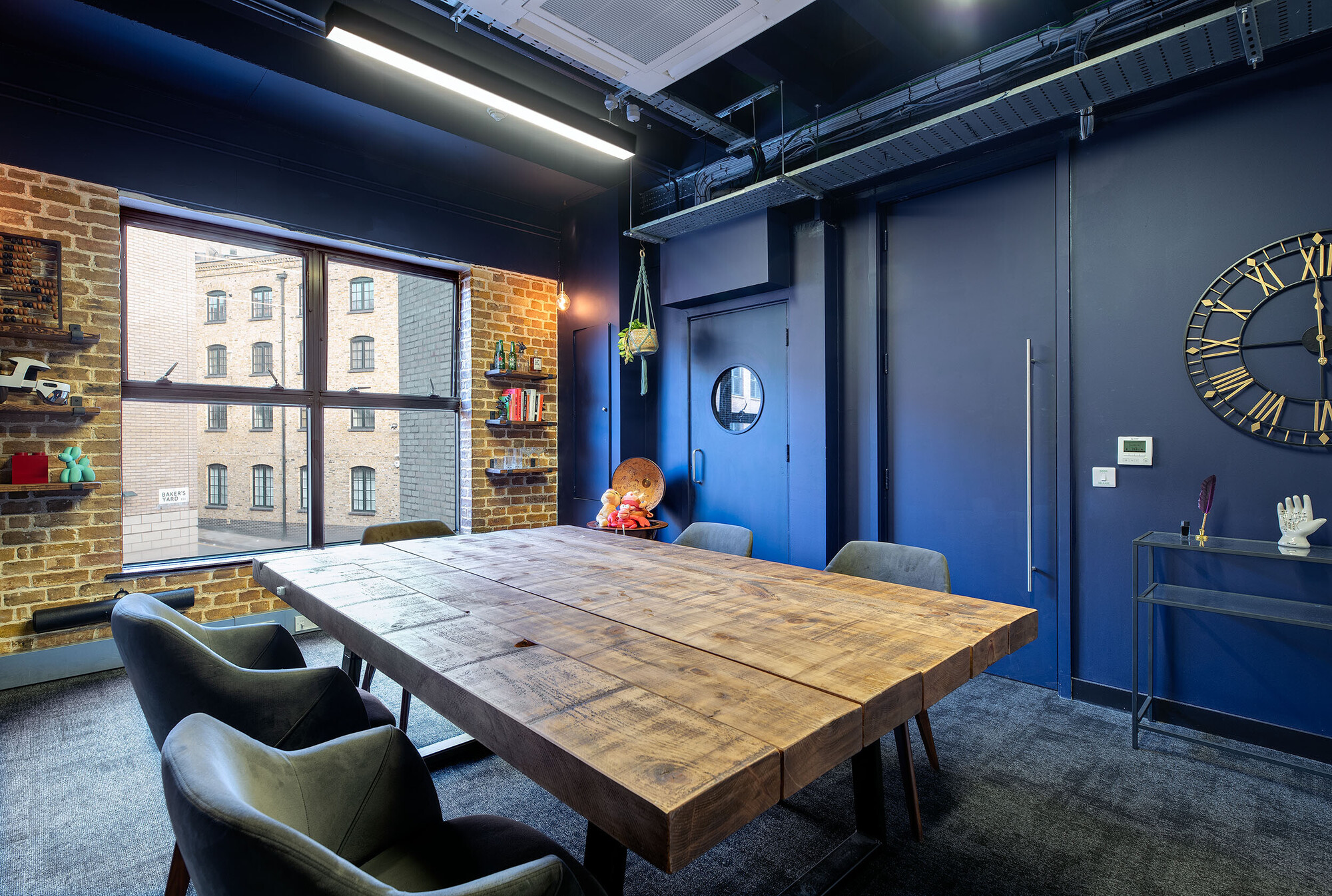 Rustic meeting room at Wiser designed by Two, featuring a large, reclaimed wood table, industrial shelving, and navy walls with brick accents.