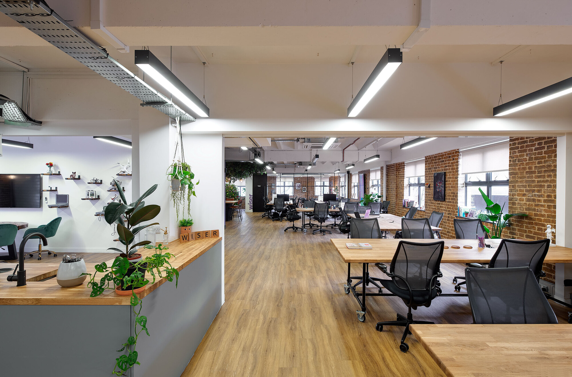 An open-plan workspace designed by Two for Wiser, featuring brick walls, wooden desks, and a greenery-filled reception area.