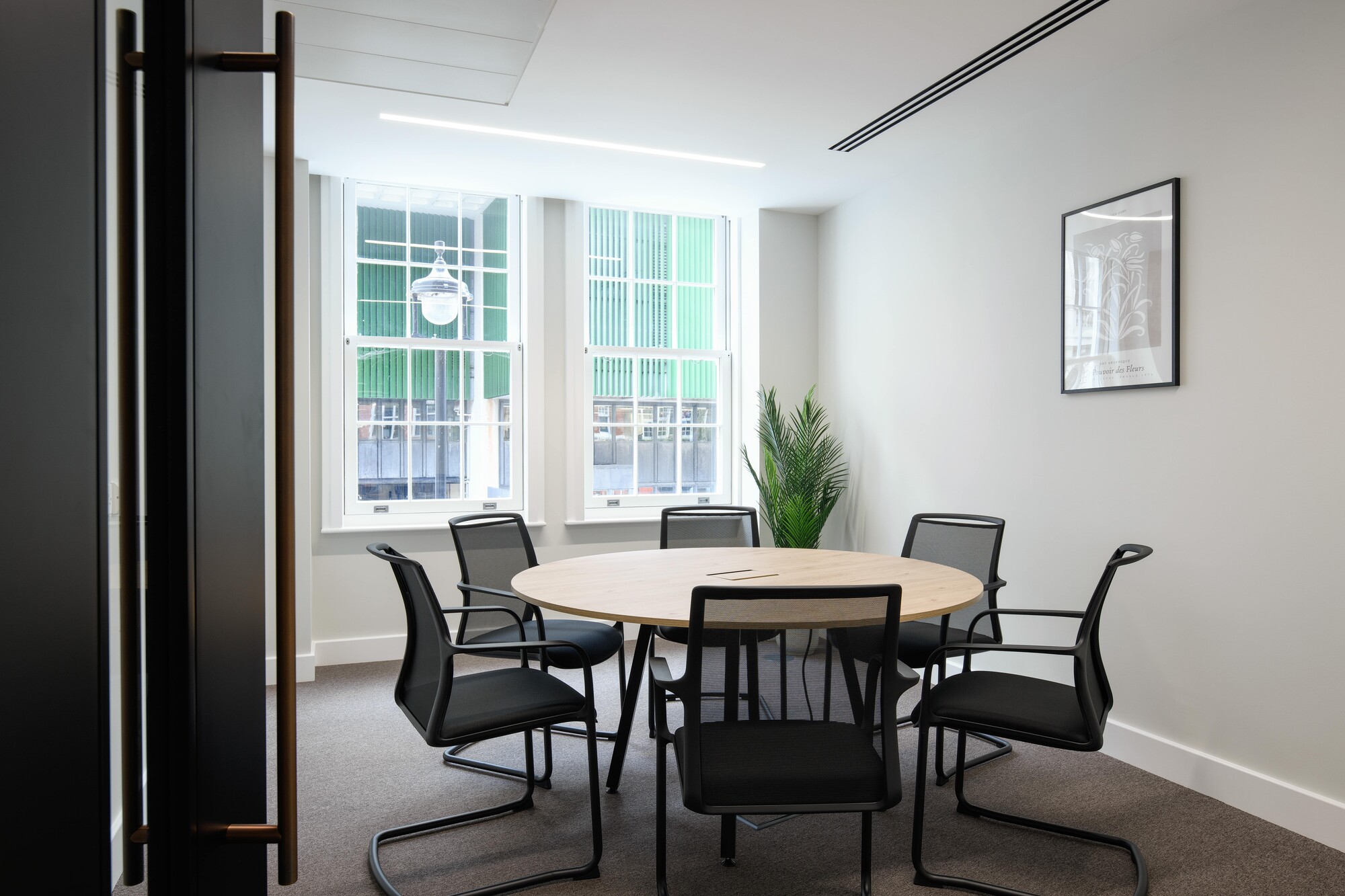 Compact meeting room designed by Two for an Asset Management Firm, featuring a round wooden table, sleek black chairs, and abundant natural light from large windows.