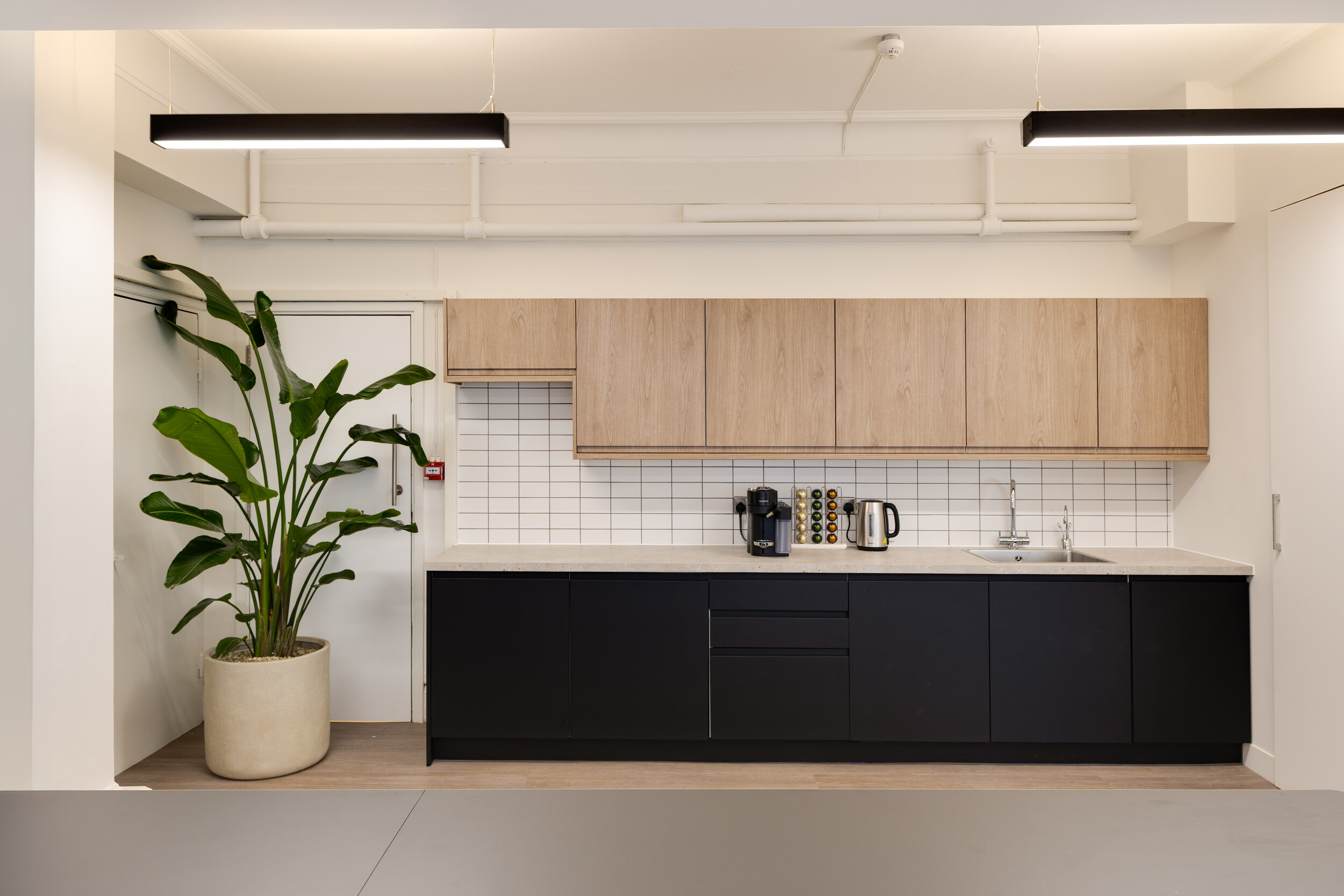 Modern InPost teapoint with black lower cabinets, light wood upper cupboards, white tiled backsplash, coffee machine, kettle, and a tall potted plant.