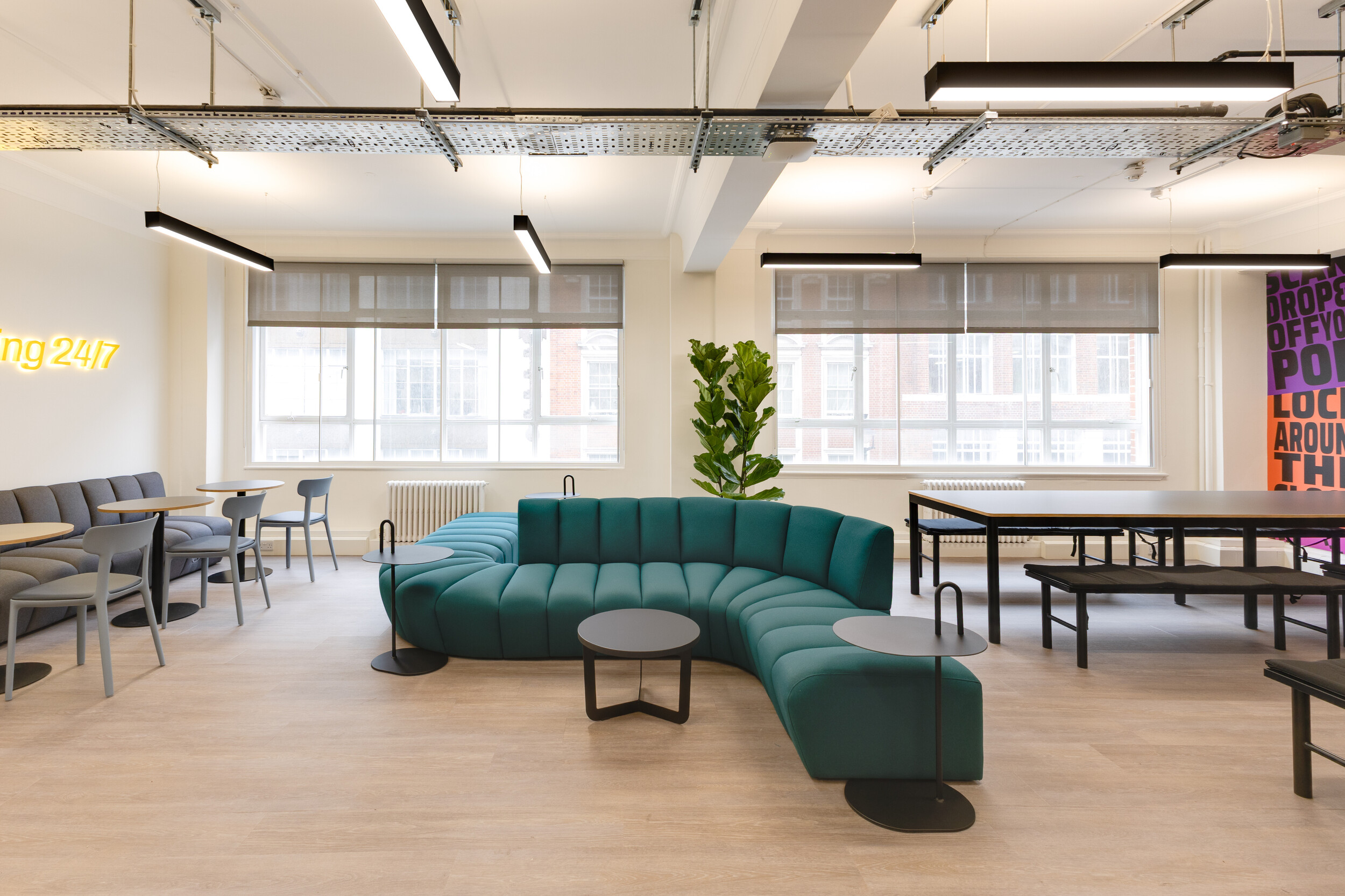 InPost office breakout space with curved teal seating, black side tables, grey dining chairs, large windows, and a leafy indoor plant.