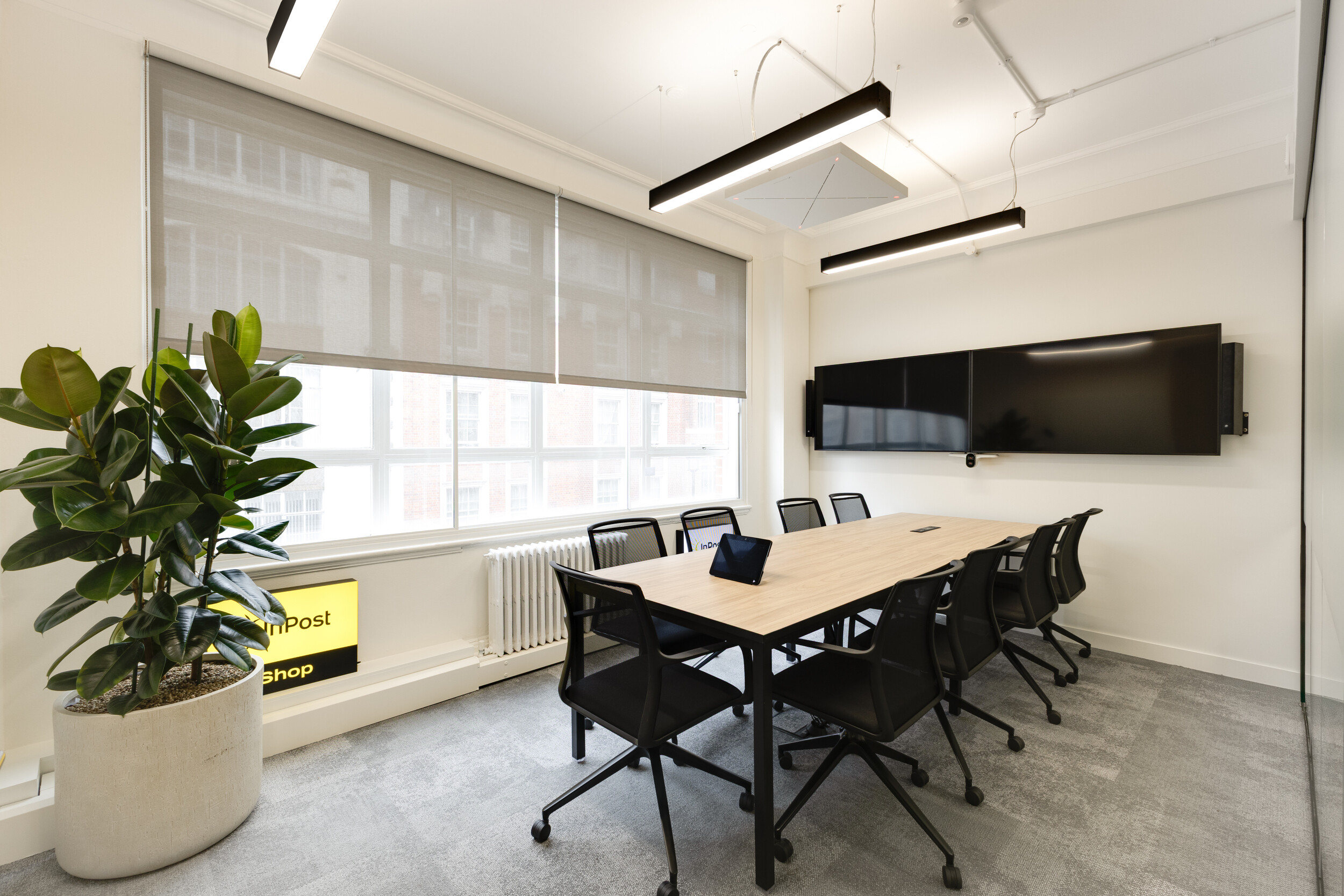 InPost conference room with wooden table, black mesh chairs, dual wall-mounted screens, potted plant, and large window with grey blinds.