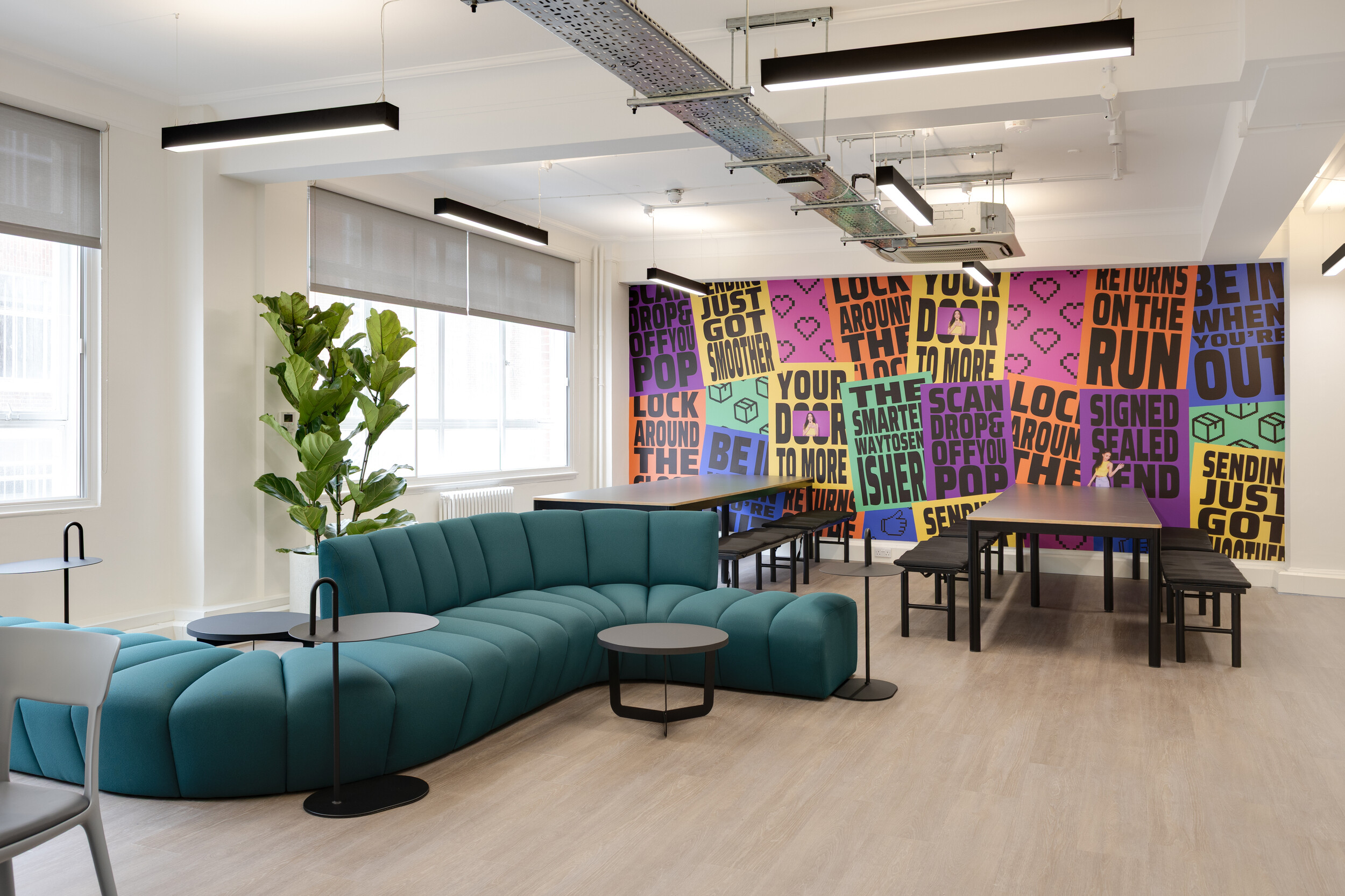 InPost breakout space with curved teal sofa, black side tables, communal dining tables, and a vibrant feature wall with bold parcel-themed graphics.