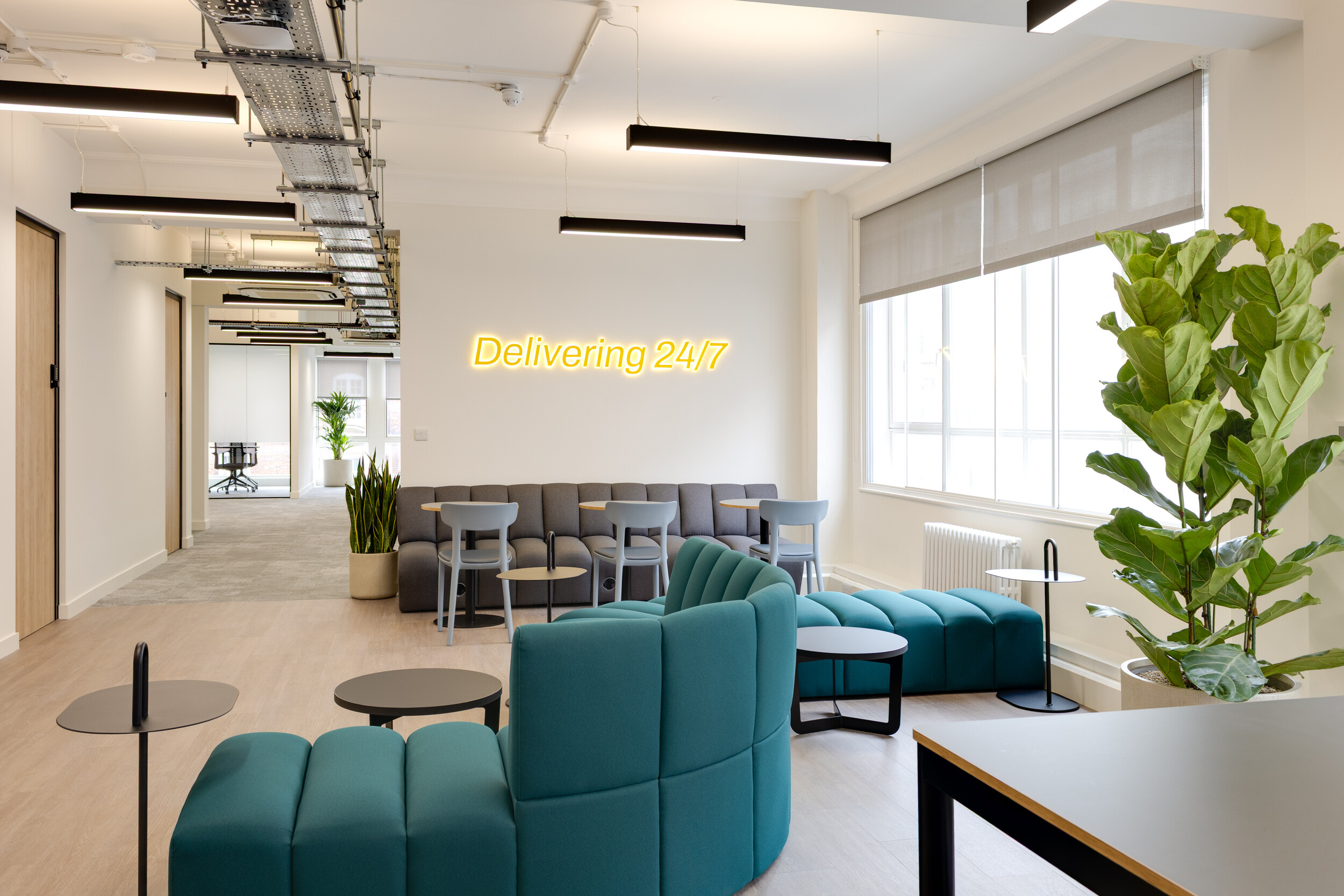InPost office lounge with teal curved seating, grey chairs, large potted plants, and a neon yellow “Delivering 24/7” sign on the wall.