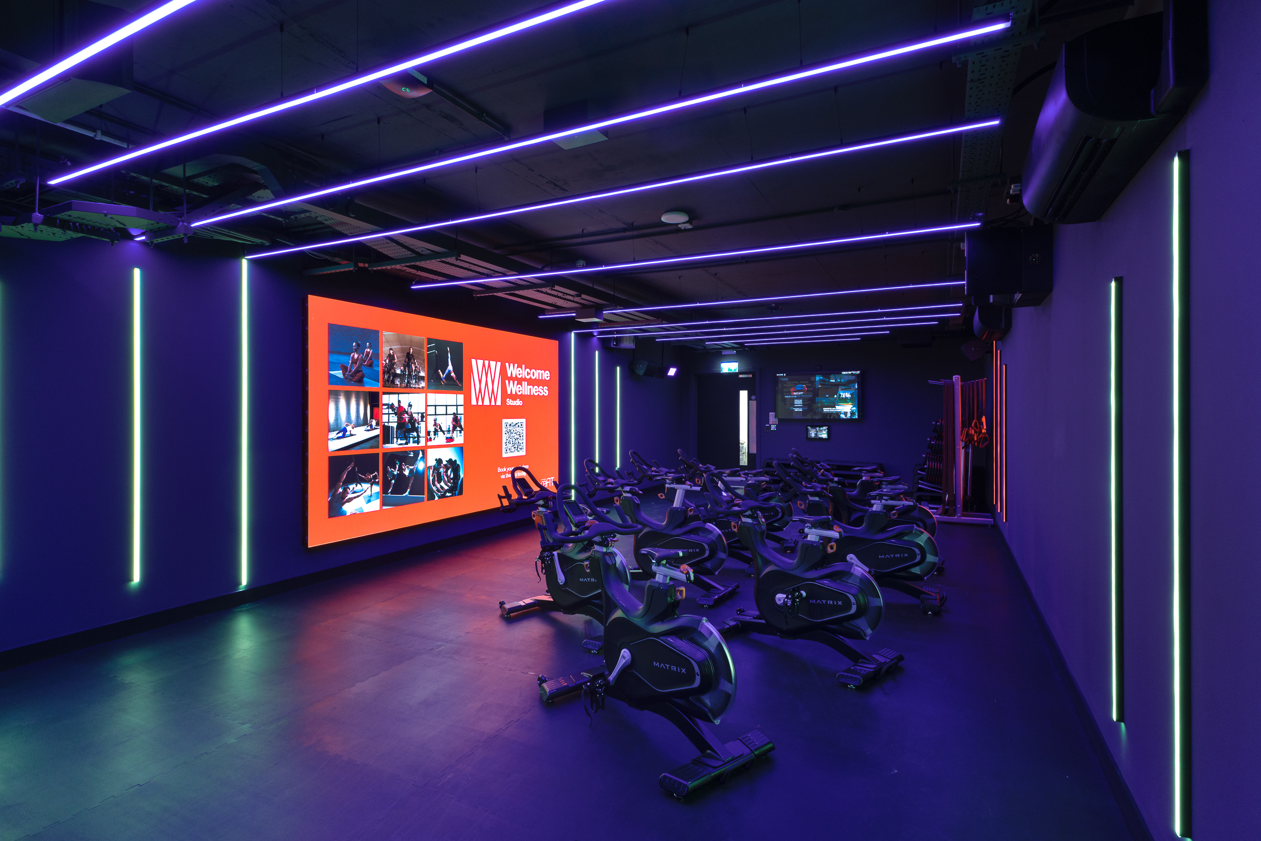 Indoor cycling studio at reFIT Bristol, the Welcome Building, with Matrix bikes, neon ceiling lights, and a bright red Welcome Wellness display screen created for energetic group workouts.