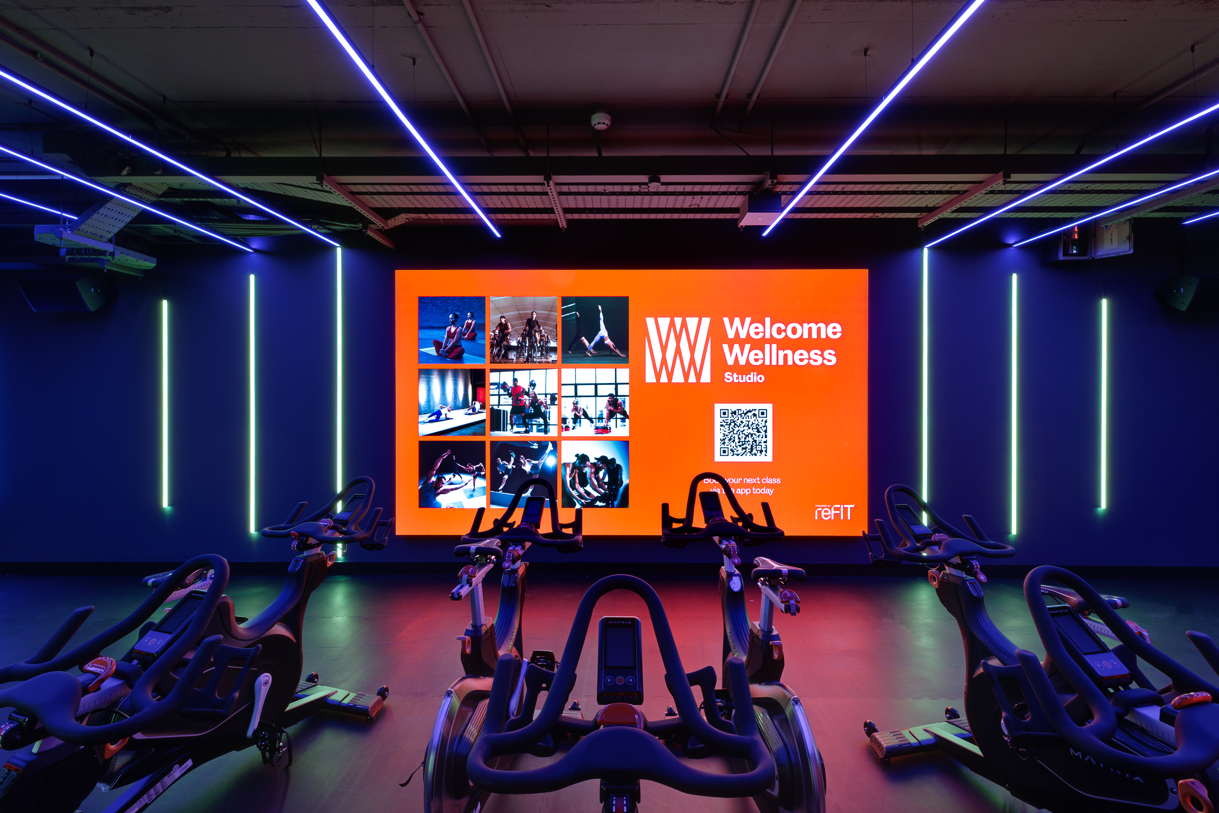 Close-up of the cycling studio at reFIT Bristol, the Welcome Building, showing Matrix bikes facing a large red Welcome Wellness screen under neon blue and green lighting.