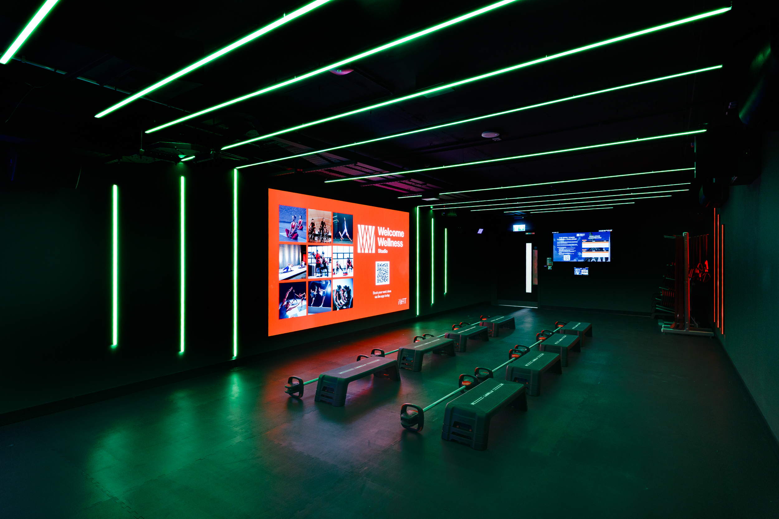 Workout studio at reFIT Bristol, the Welcome Building, featuring step benches and kettlebells arranged under green neon lighting with a red Welcome Wellness display at the front.