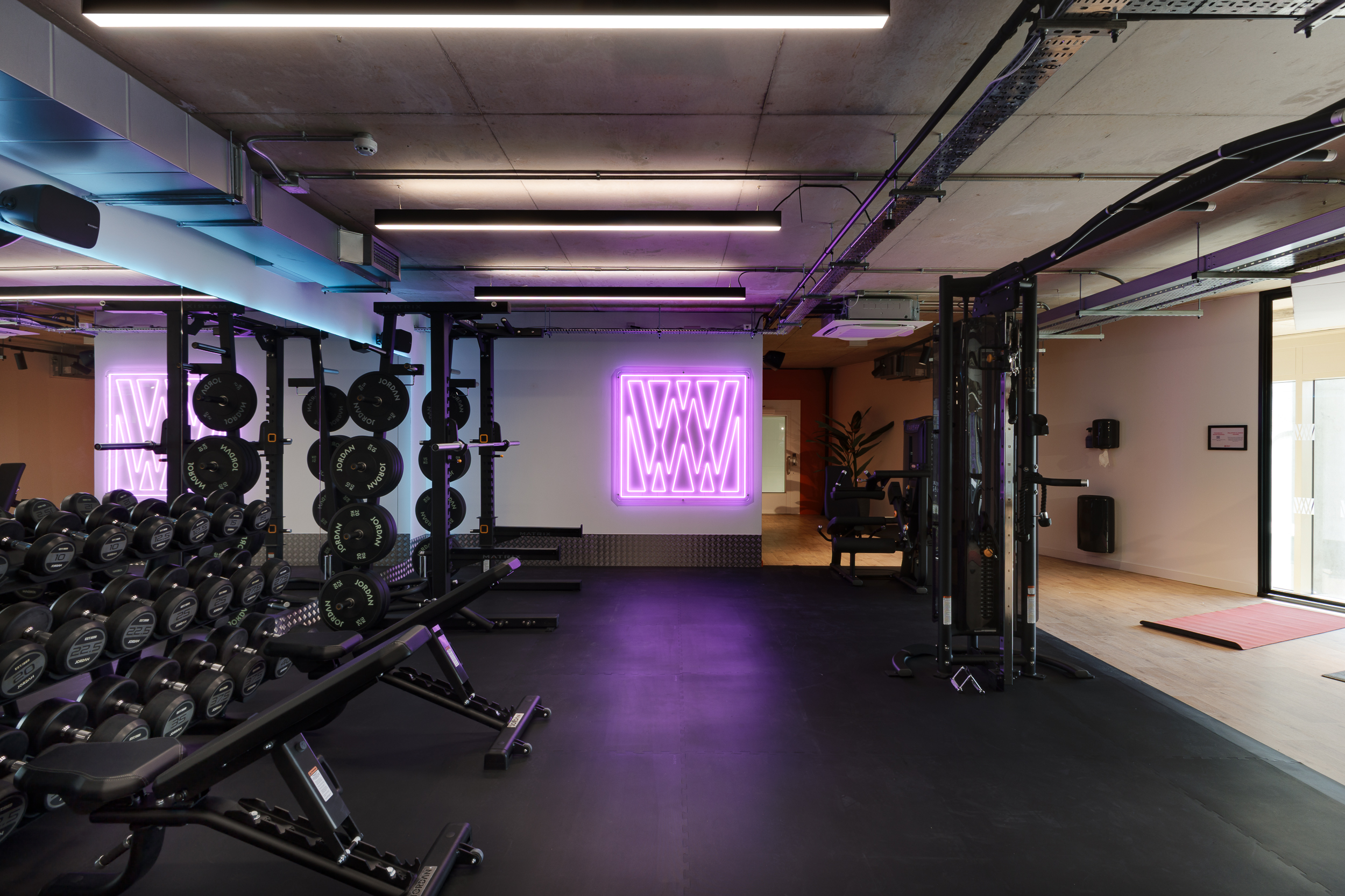 Gym area at reFIT Bristol, the Welcome Building, with weightlifting racks, dumbbells, and a purple illuminated ‘W’ wall sign beside an open workout zone.