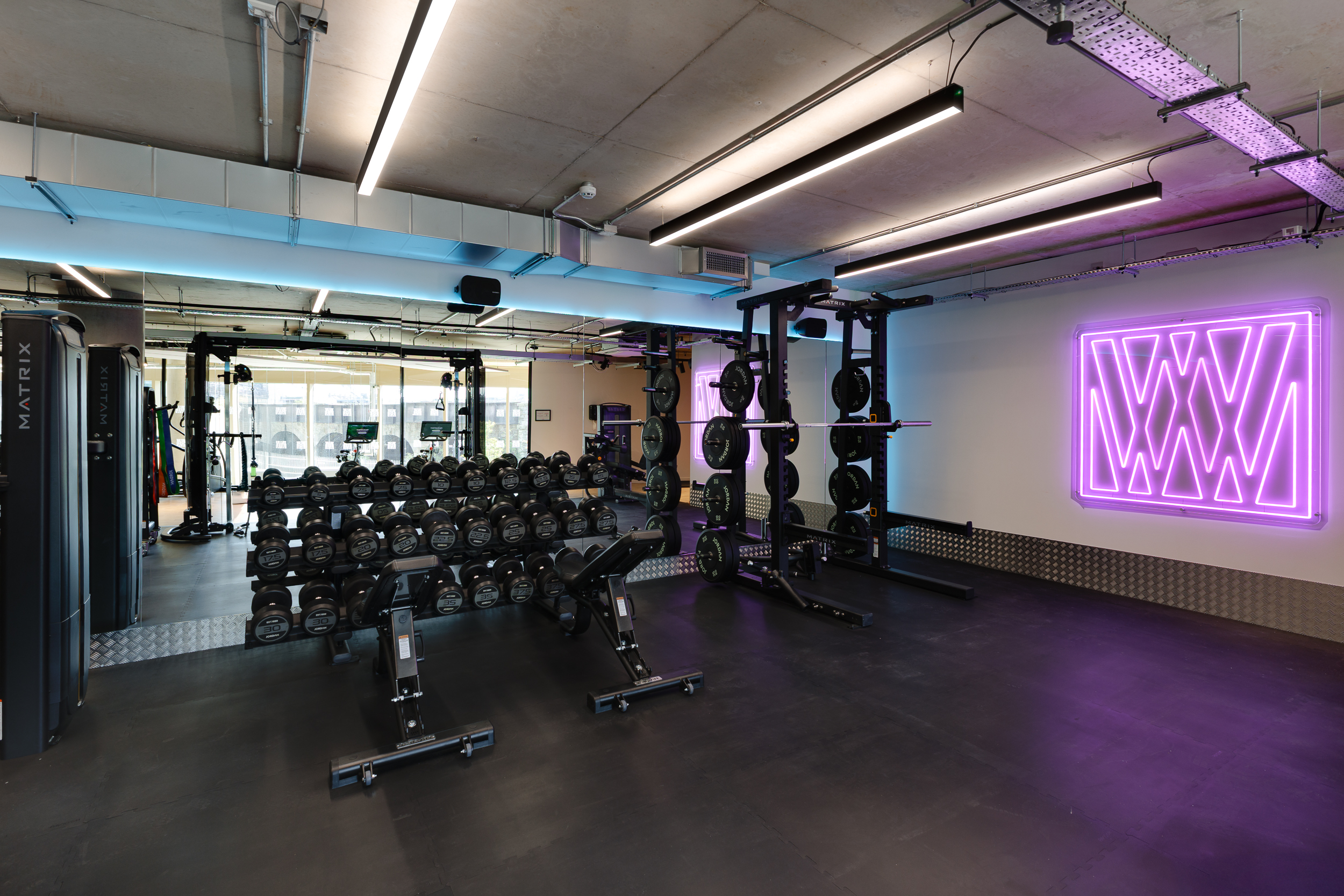 Modern gym at reFIT Bristol, the Welcome Building, featuring dumbbell racks, squat racks, and a glowing purple ‘W’ light on the wall beside large windows.